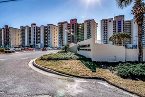 A multi-story residential building with a curved road in front, a palm tree, and a flag. - Casual beachfront getaway with pool, ocean views, & beach access (Orange Beach)