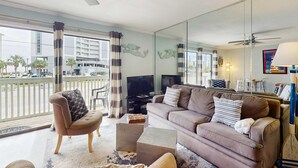 A living room with a sofa, armchair, TV, and a balcony with a view of buildings and palm trees. - Coastal condo close to the beach with balcony - walk everywhere! (Gulf Shores)