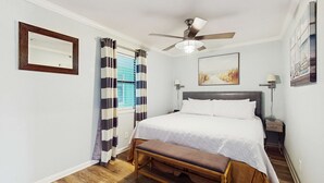 A bedroom with a bed, a ceiling fan, a bedside table, a lamp, a window with curtains, and a wall-mounted mirror. - Coastal condo close to the beach with balcony - walk everywhere! (Gulf Shores)