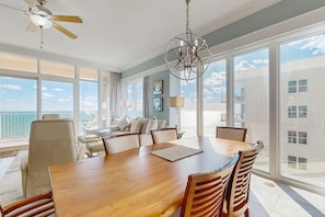 A spacious dining area with a large wooden table and chairs, a ceiling fan, and large windows offering a view of the ocean. - Gulf-front condo with panoramic views, balcony & shared pool, hot tub, gym (Gulf Shores)