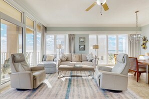 A spacious living room with a large window, a ceiling fan, and a dining area. - Gulf-front condo with panoramic views, balcony & shared pool, hot tub, gym (Gulf Shores)