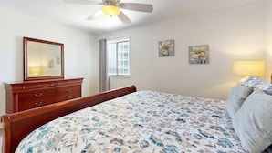 Un dormitorio con cama, ventilador de techo, cómoda, ventana y dos cuadros de flores en la pared.