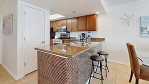 Una cocina con armarios de madera, encimera de granito, microondas y un lavabo. Hay dos taburetes y una mesa con una silla.