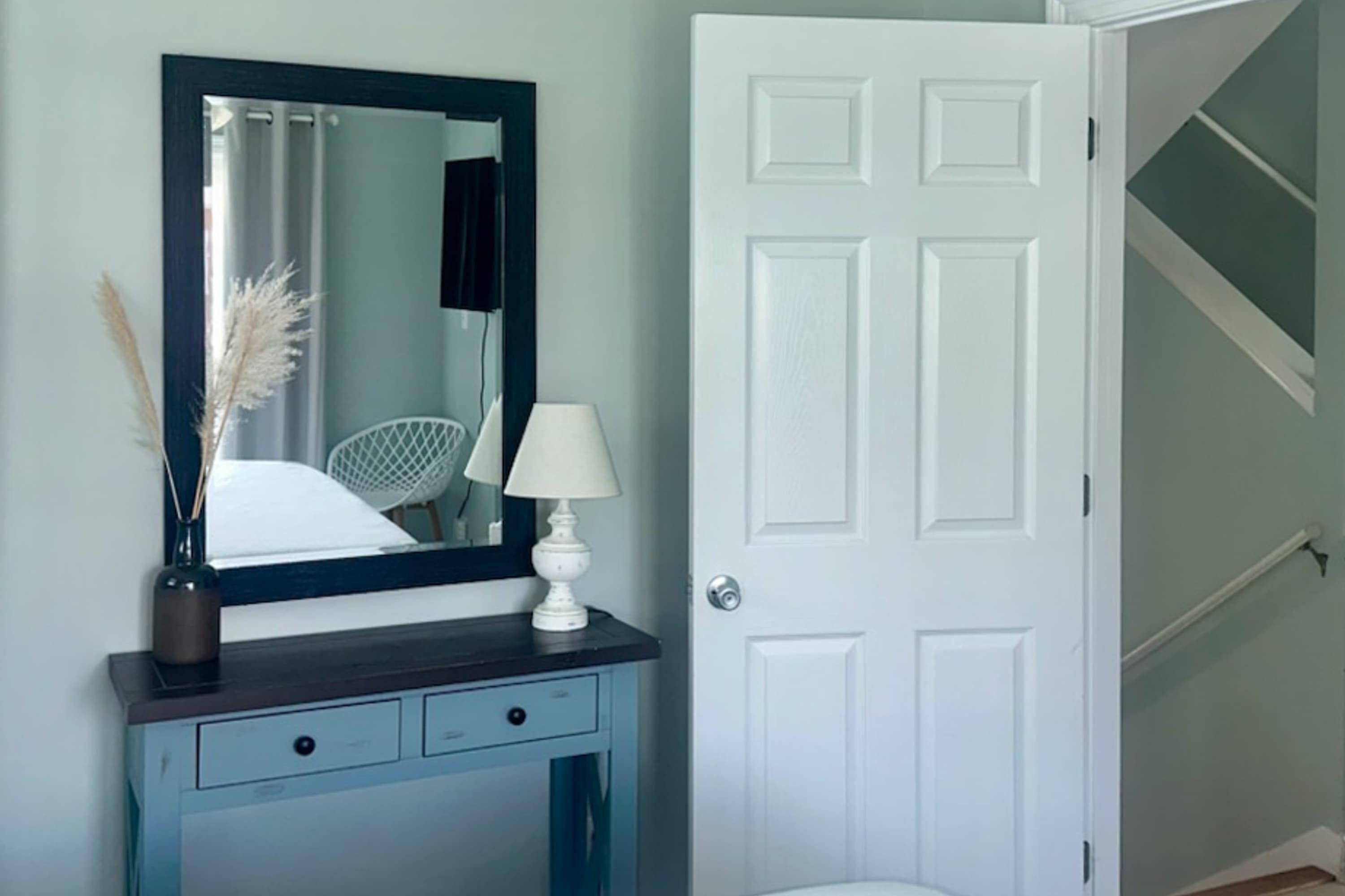 A room with a dark blue vanity, a white door, and a mirror reflecting a bed and a lamp.