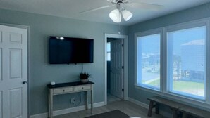 Un dormitorio con cama, televisor montado en la pared, ventilador de techo, ventana con persianas y puerta que da a otra habitación.