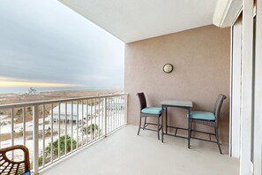 A balcony with a view of the beach, two wicker chairs, a small table, and a wall-mounted light fixture. - The Dunes 603 | Beachside Condo with Gulf Views and Swim-Through Pool (Gulf Shores)