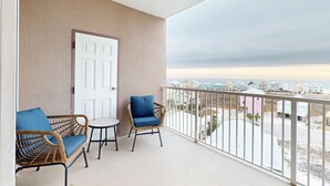 Un balcón con mobiliario de mimbre, una puerta blanca y vistas a edificios.