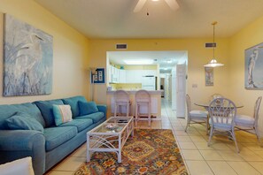 A living room with a sofa, coffee table, and chairs, a kitchen in the background, and a dining area with a table and chairs. - The Dunes 603 | Beachside Condo with Gulf Views and Swim-Through Pool (Gulf Shores)