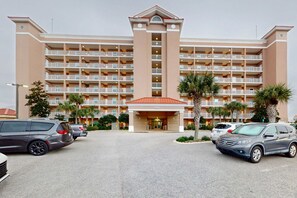 Een meerlaags hotel met een centrale ingang, balkons op elke verdieping en een parkeerplaats met verschillende auto's.