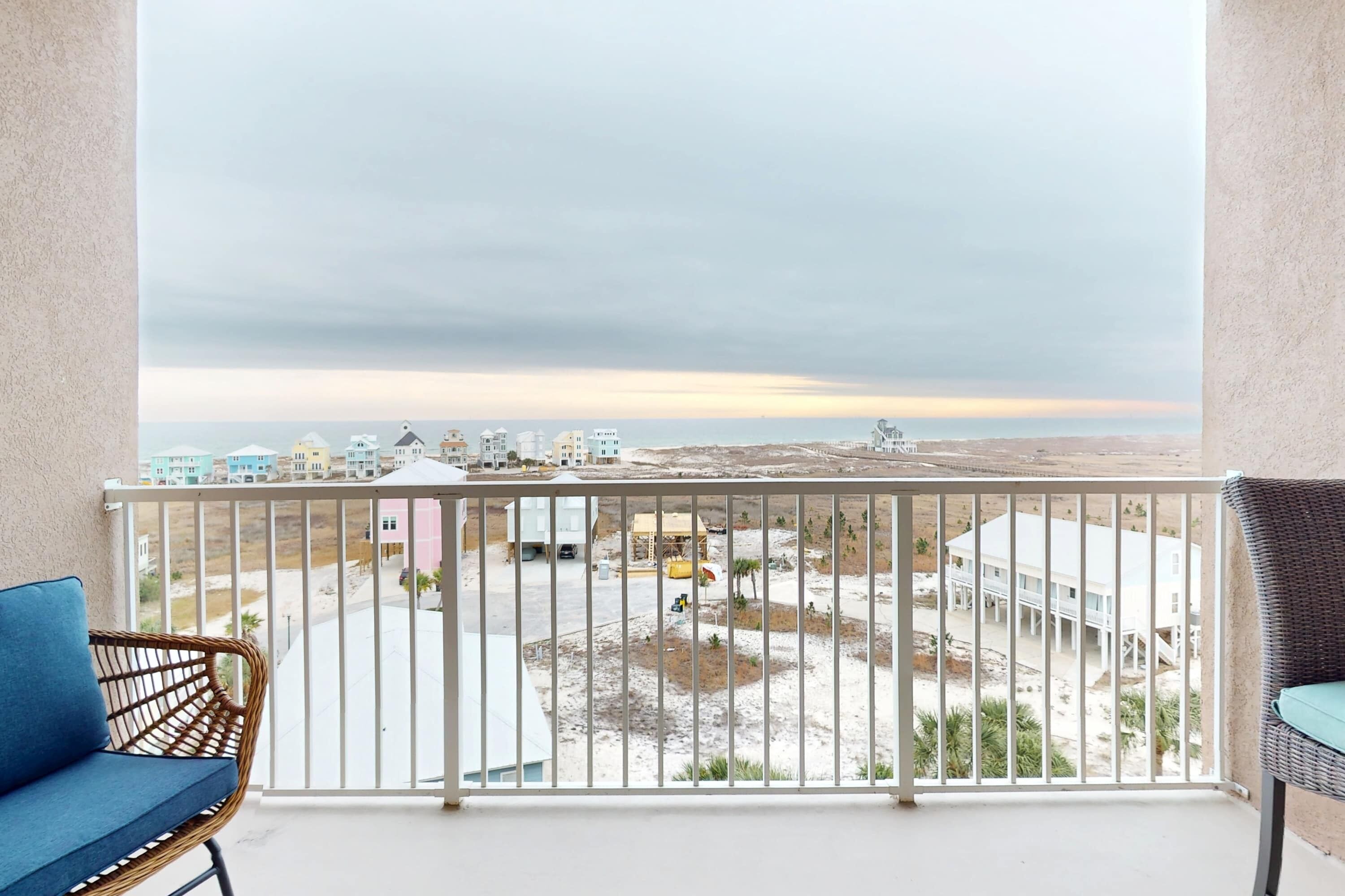 Un balcón con vista a la playa y edificios, una silla de mimbre y una barandilla blanca.
