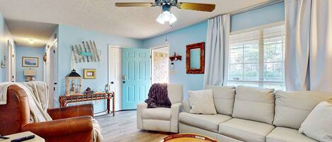 A living room with a white sofa, a brown leather chair, a ceiling fan, and a wicker coffee table.