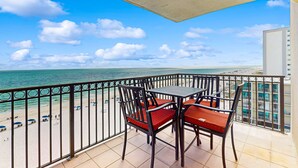 A balcony with a view of the beach and ocean, featuring a table and chairs. - Beachfront condo with shared pool, outdoor shower, and beach access (Gulf Shores)