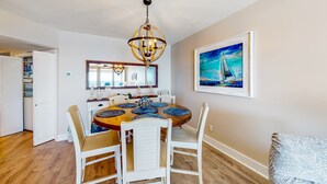 A dining area with a round table, white chairs, a chandelier, and a sailboat painting on the wall. - Dreamy beach condo with water views, balcony, pool & beach access (Orange Beach)