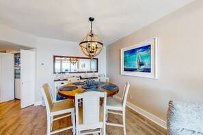A dining area with a round table, white chairs, a chandelier, and a sailboat painting on the wall. - Dreamy beach condo with water views, balcony, pool & beach access (Orange Beach)
