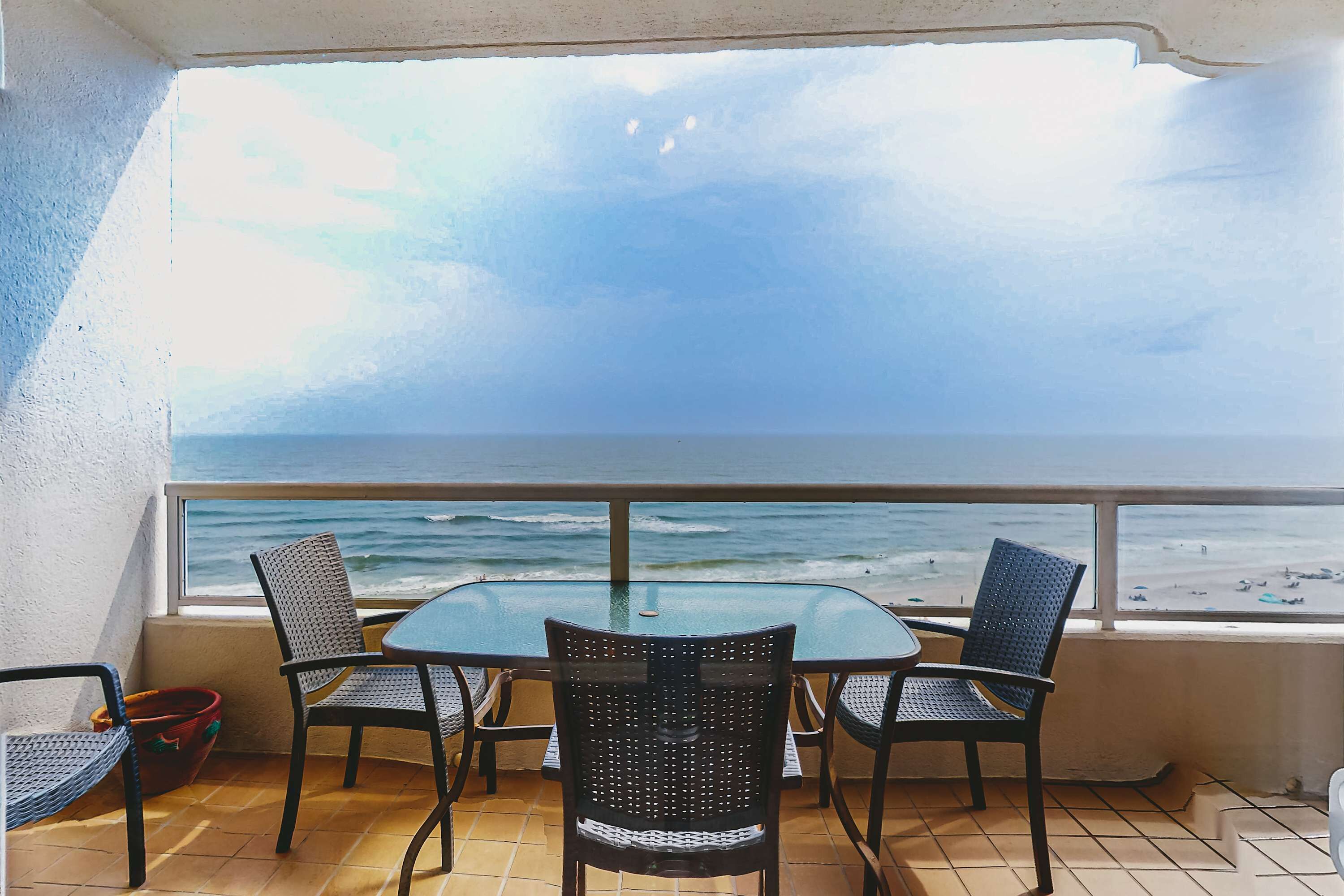 A balcony with a glass table and four chairs, overlooking the ocean.