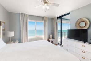 A hotel room with a bed, a TV, a ceiling fan, a window with curtains, and a view of the ocean. - Fantastic Home Located in Beachfront Building w/Shared Pool & Hot Tub, WiFi, AC (Orange Beach)