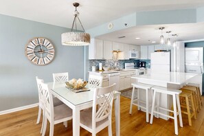 A modern kitchen with white cabinetry, a central island, and a dining area with white chairs and a glass table. - Fantastic Home Located in Beachfront Building w/Shared Pool & Hot Tub, WiFi, AC (Orange Beach)