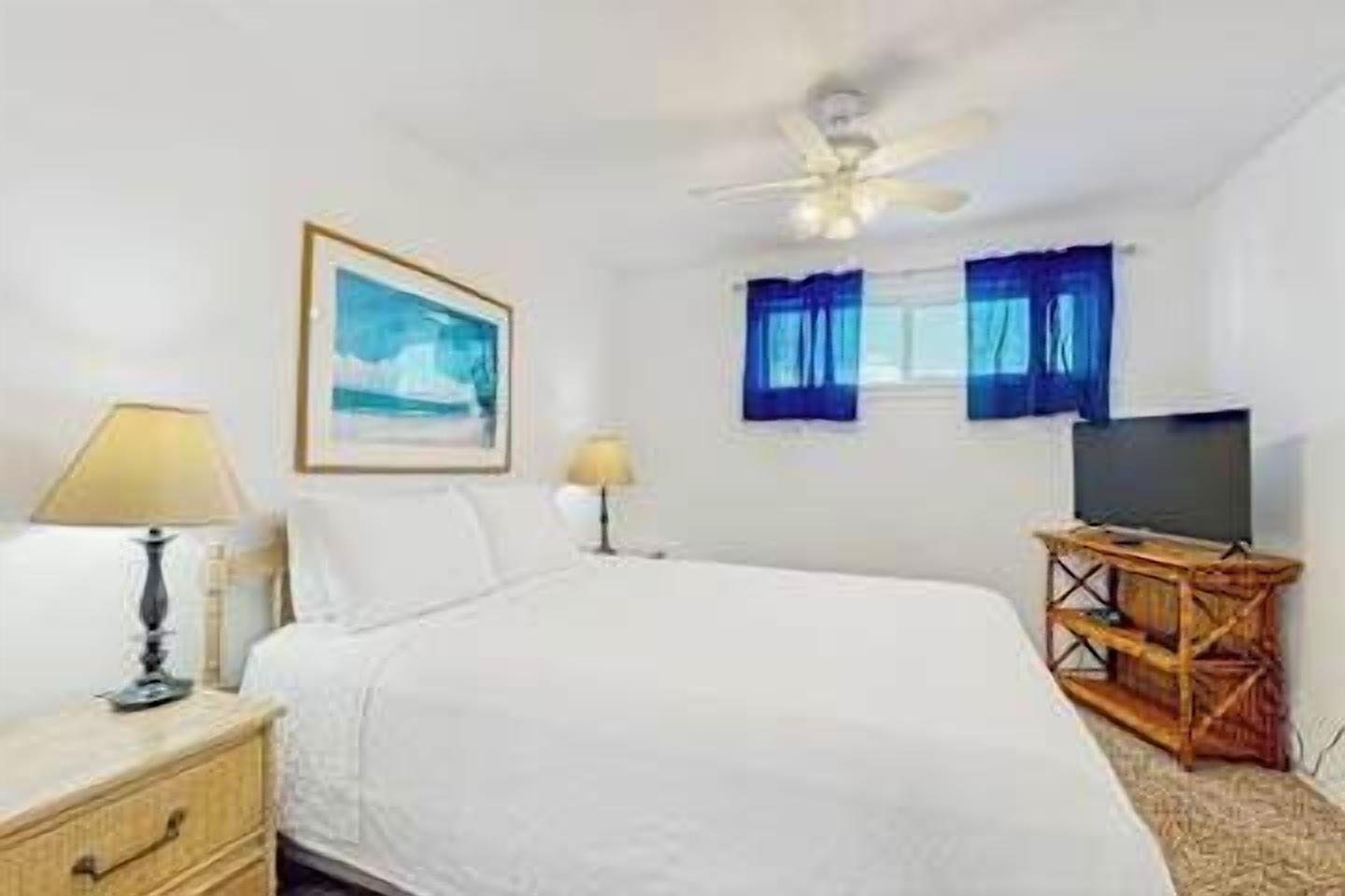 A hotel room with a bed, a television on a wooden stand, a ceiling fan, and a window with curtains.