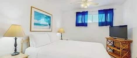 Habitación de hotel con cama, televisor sobre un soporte de madera, ventilador de techo y ventana con cortinas.