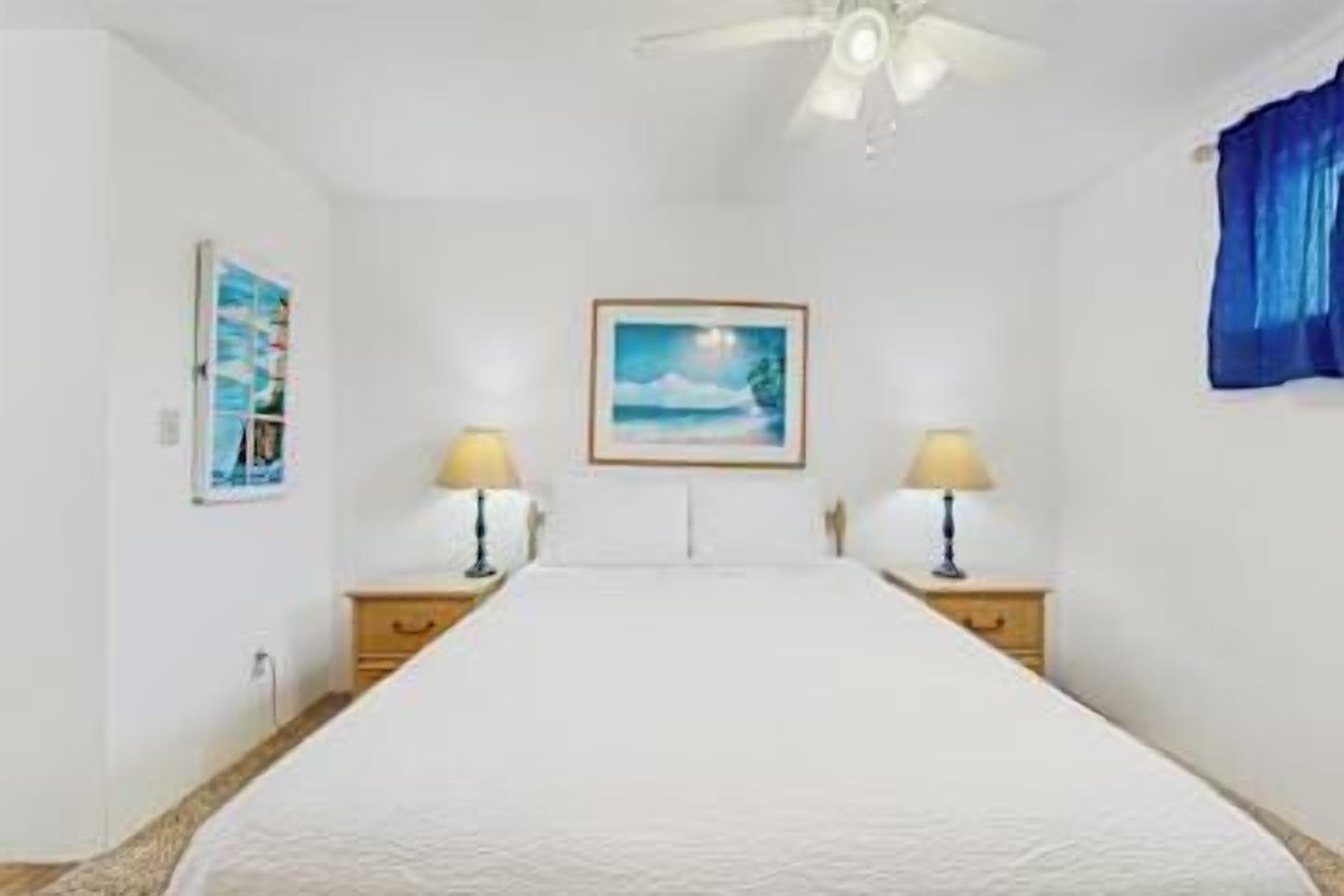 A bedroom with a bed, two bedside tables with lamps, a ceiling fan, and a framed picture on the wall.