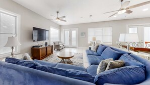Ein geräumiges Wohnzimmer mit einem großen, blauen Sofa, einem Holztisch und einem Flachbildfernseher. Es gibt Deckenventilatoren, eine Holzkommode und einen Essbereich mit Tisch und Stühlen.