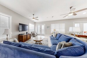 Un salon spacieux avec un grand canapé bleu, une table basse en bois et un téléviseur à écran plat. On y trouve également des ventilateurs de plafond, un meuble en bois et un coin repas avec une table et des chaises.