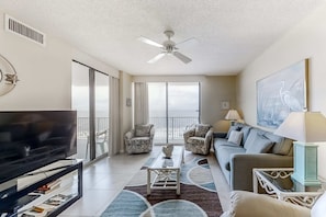 A living room with a TV, sofa, chairs, a ceiling fan, and a balcony with a view. - Fun, beachy condo in Gulf-front complex with pool & tennis court (Orange Beach)