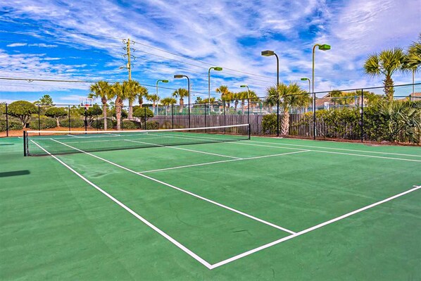 A tennis court with a net, surrounded by a fence, palm trees, and street lamps. - Beachfront condo with pool, tennis court, hot tub & central AC (Orange Beach)