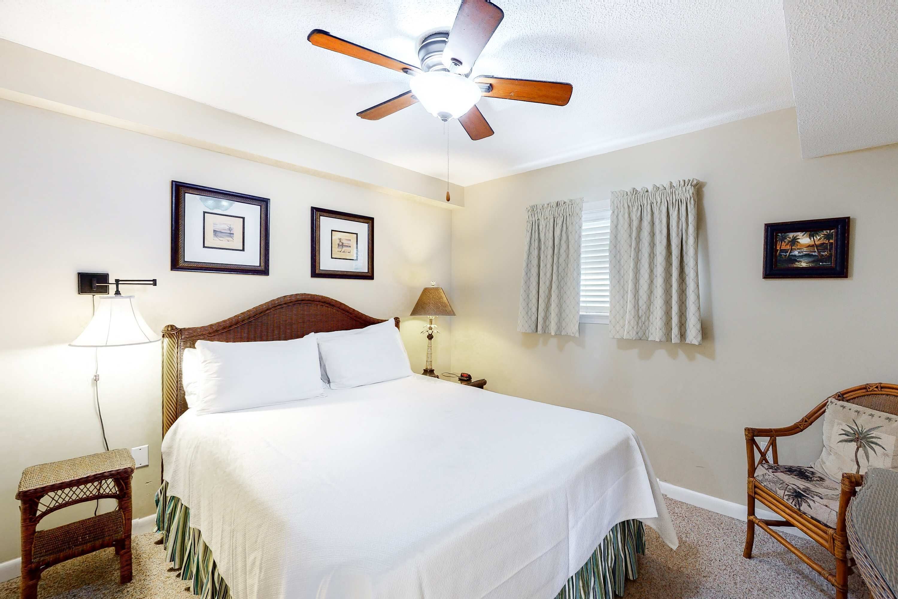 A bedroom with a bed, a ceiling fan, a nightstand, a lamp, a chair, and two framed pictures on the wall.