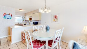 A dining area with a round glass table, white chairs, a white bar, and a kitchen in the background. - Gorgeous home in beachfront complex with pool, tennis court & WiFi (Orange Beach)