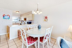 A dining area with a round glass table, white chairs, a white bar, and a kitchen in the background. - Gorgeous home in beachfront complex with pool, tennis court & WiFi (Orange Beach)