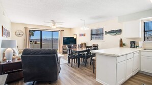 Une cuisine moderne avec des meubles blancs, un coin repas avec des chaises foncées, une grande fenêtre avec des stores et un ventilateur de plafond.