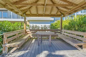 Um deck de madeira com uma mesa redonda e bancos, rodeado por vegetação e um edifício ao fundo.