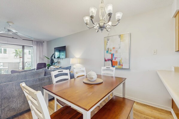 A dining area with a wooden table, white chairs, a chandelier, and a large abstract painting on the wall. - Wonderful Beachside Home with Free WiFi, Central AC, Shared Pool, & Elevator (Orange Beach)