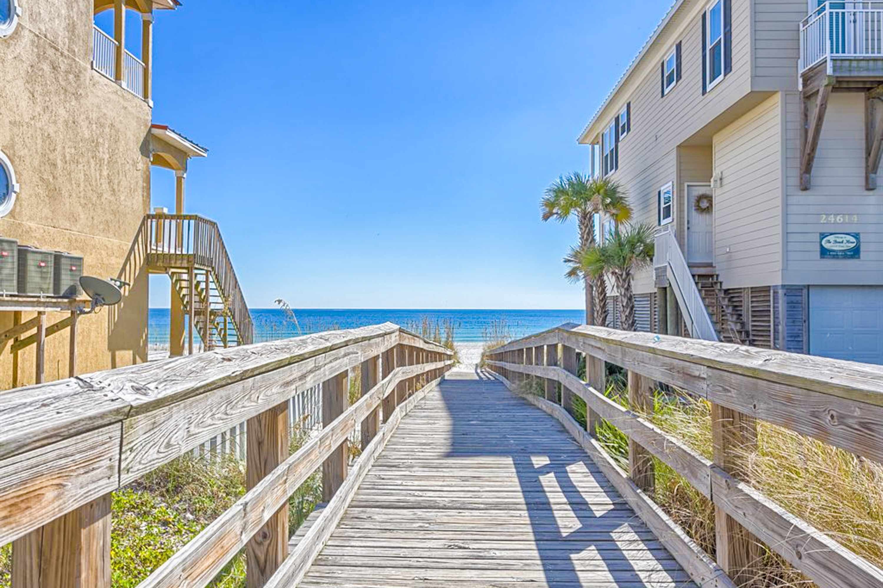 A wooden boardwalk leading to a beach, flanked by buildings with balconies and a clear view of the ocean.