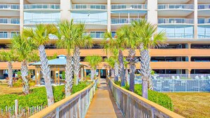 A multi-story hotel with a central walkway, palm trees, and outdoor seating areas. - Gulf-front condo with shaded balcony, pools, hot tubs, gym & beach access (Orange Beach)
