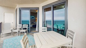 Un balcon avec une table et des chaises, une porte-fenêtre coulissante donnant sur une pièce avec vue sur l’océan, et un ventilateur de plafond.