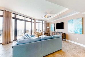 Un salon spacieux avec un grand canapé d’angle, un ventilateur de plafond, un téléviseur à écran plat et des œuvres d’art accrochées aux murs.