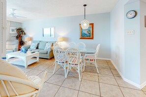 A living room with a sofa, coffee table, dining table with chairs, a hanging light fixture, a clock, and a framed picture on the wall. - Beachfront condo with Gulf views, balcony, pool, hot tub, sauna & gym (Orange Beach)