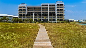 A multi-story hotel with a central courtyard, a wooden boardwalk leading to the entrance, and a clear blue sky. - Gulf-front condo with inviting balcony, amazing views, pool & hot tub (Orange Beach)