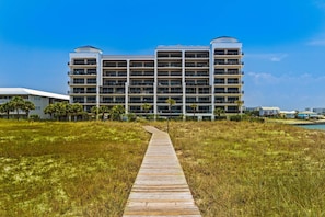 A multi-story hotel with a central courtyard, a wooden boardwalk leading to the entrance, and a clear blue sky. - Gulf-front condo with inviting balcony, amazing views, pool & hot tub (Orange Beach)