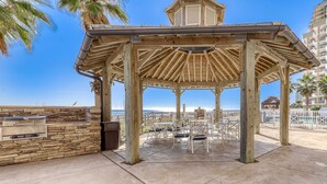 A wooden gazebo with stone walls, outdoor seating, and a view of the ocean. - Convenient oceanfront condo with views, balcony, pool, hot tub, A/C, & WiFi (Orange Beach)