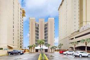 A multi-story hotel with a central entrance, parking lot, and palm trees. - Gulf & Beachfront Condo wBalcony, Central AC, Shared Hot Tub, Pools, Free WiFi (Orange Beach)