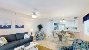 Una sala de estar con sofá, mesa de centro y sillón estampado, un comedor con sillas y mesa, y una cocina al fondo.