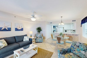 A living room with a sofa, coffee table, and patterned armchair, a dining area with chairs and a table, and a kitchen in the background. - Gulf & Beachfront Condo wBalcony, Central AC, Shared Hot Tub, Pools, Free WiFi (Orange Beach)