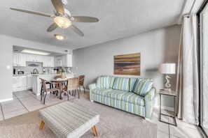 A modern living room with a sofa, a dining area with chairs, a ceiling fan, and a kitchen in the background. - Gulf front condo with great views, shared pools & beach access (Orange Beach)