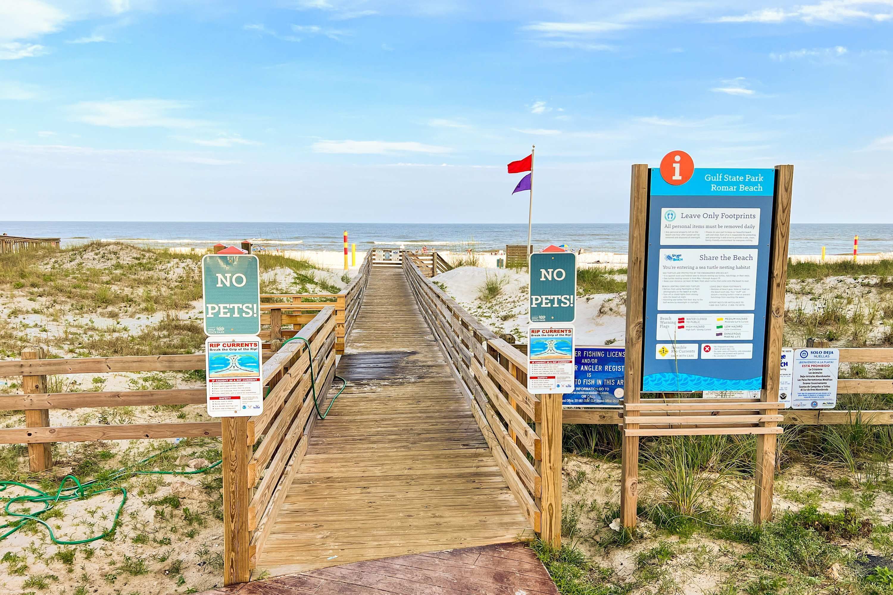 A wooden boardwalk leading to a beach with signs indicating no pets and rip currents warnings.