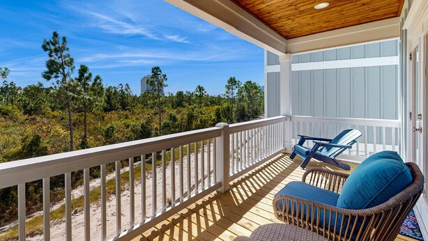 A wooden deck with a railing, a wicker chair with a cushion, and a view of the beach and trees. - Brilliant beach house located walking distance to beach with community pool (Orange Beach)