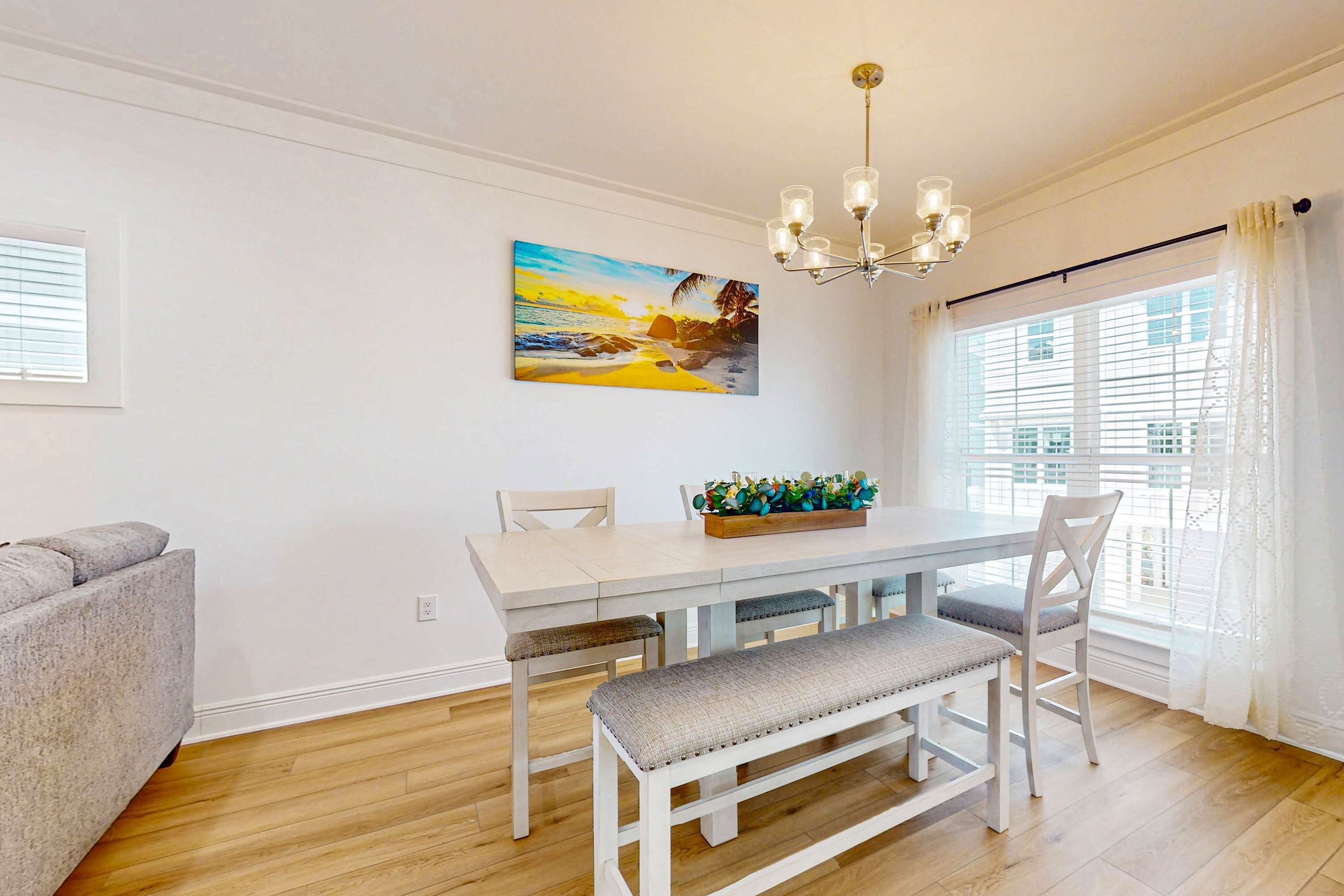 A dining area with a white table, bench, and chairs, a painting of a beach scene, and a chandelier.