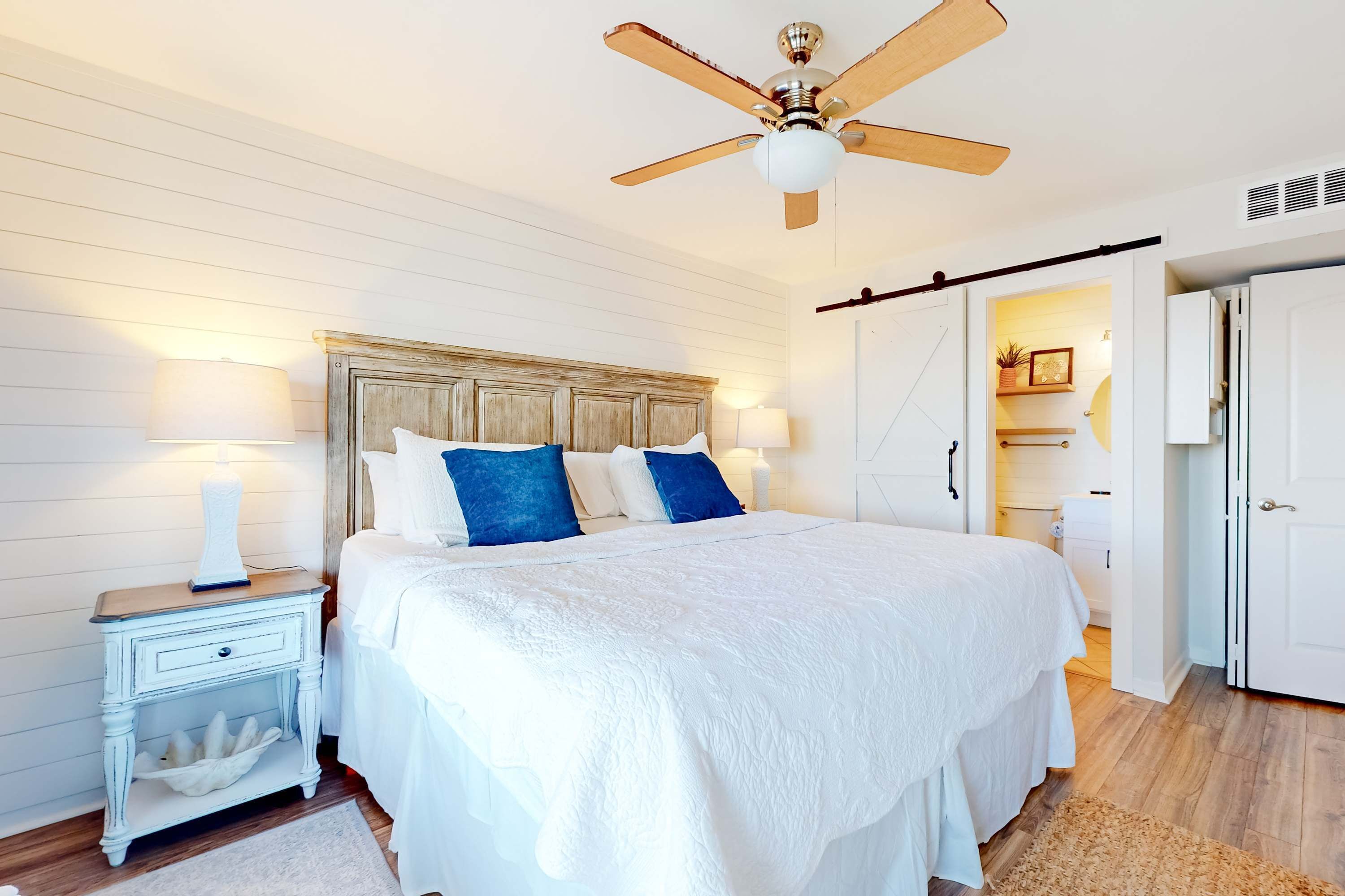 Une chambre à coucher avec une tête de lit en bois, du linge de lit blanc, une table de chevet avec une lampe et un ventilateur de plafond.
